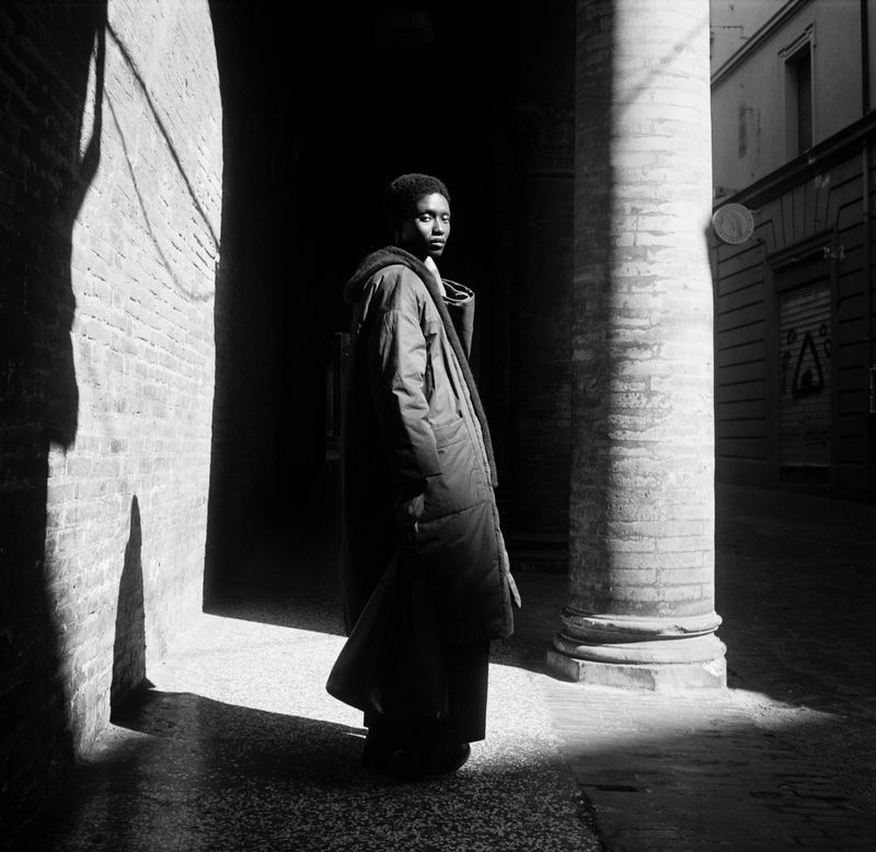 Black and white portrait of an adult male in an urban environment. The man stands partially in shadow, next to a brick pillar.