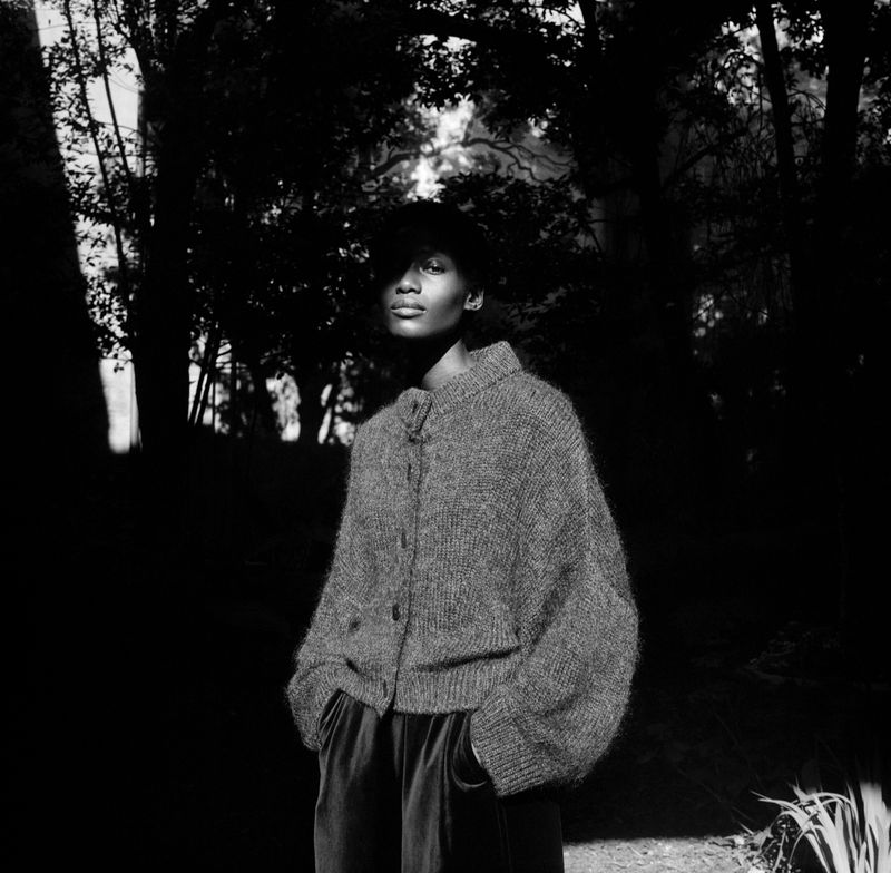 Black and white portrait of a young woman with short hair wearing a cardigan and wide-leg pants in a wooded outdoor setting.