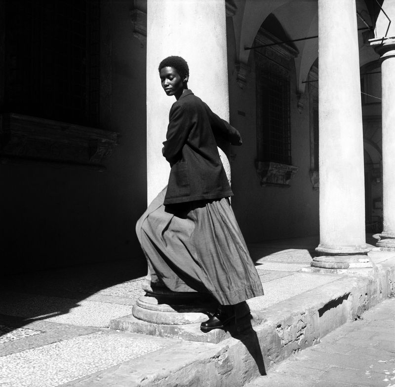 Monochrome outdoor fashion shot of an adult woman in dark blazer and long skirt, standing among a row of white columns.