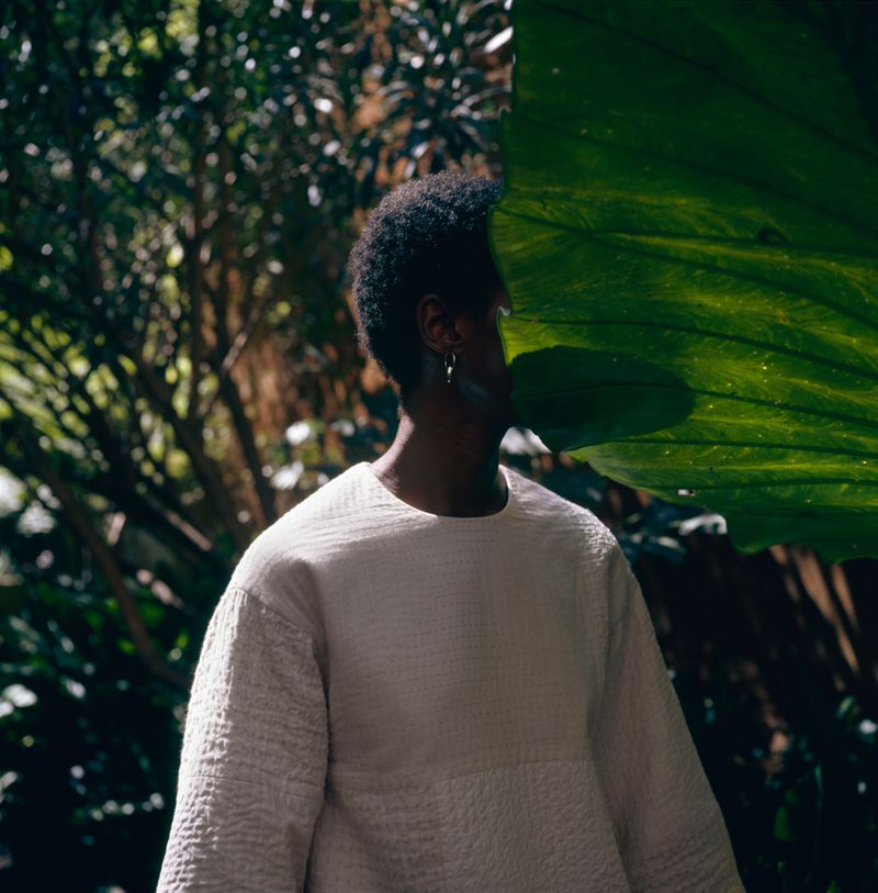 A person wearing a white textured shirt stands partially obscured by a large green tropical leaf in a lush outdoor setting.