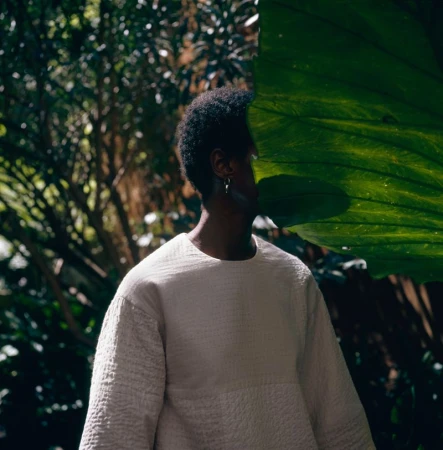 A person wearing a white textured shirt stands partially obscured by a large green tropical leaf in a lush outdoor setting.
