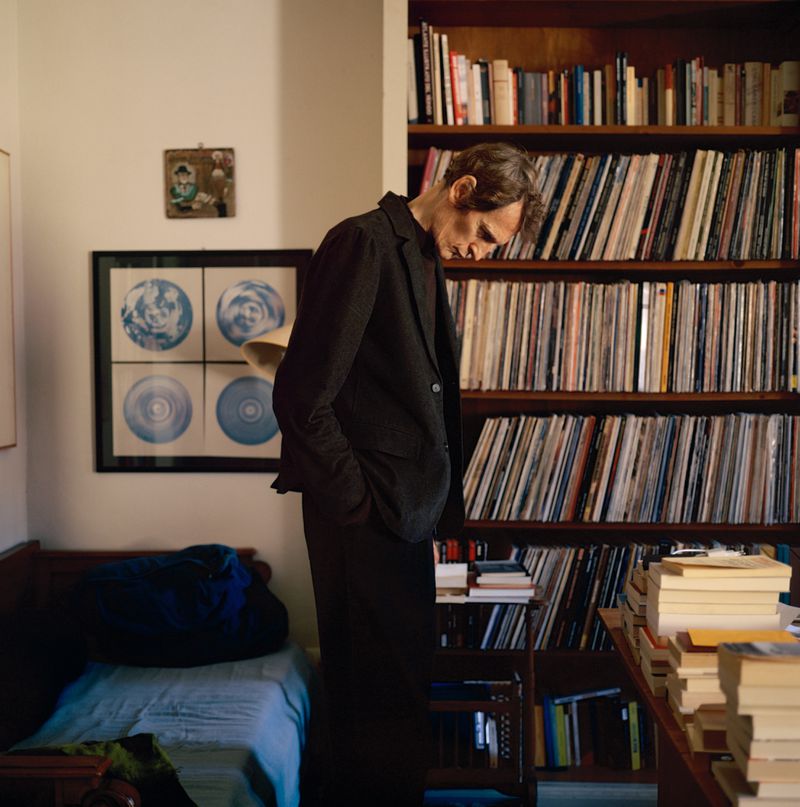 Man in dark suit standing in room with bookshelf full of records and books, warm light, indoor setting, portrait style.