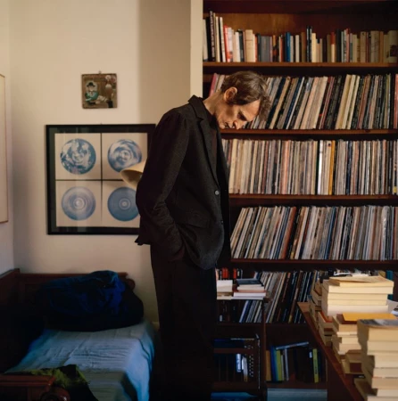 Man in dark suit standing in room with bookshelf full of records and books, warm light, indoor setting, portrait style.