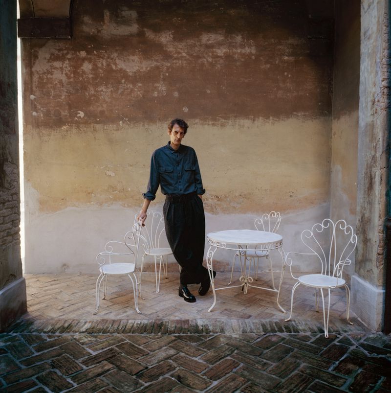 Adult man in dark shirt and pants leans against white chair on brick patio with stone wall in the background, outside on a sunny day.