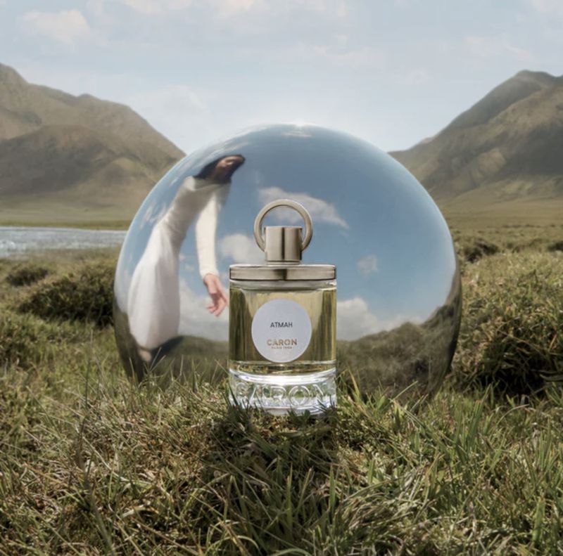 Perfume bottle in a grassy field with rolling hills in the background. The bottle is partially reflected in a spherical object.