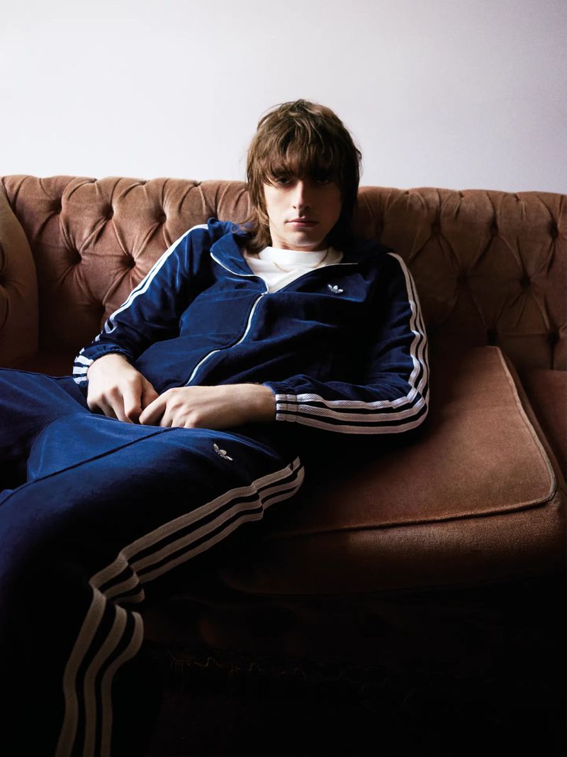 Man lounges on a brown tufted couch wearing a blue tracksuit with white stripes, looking directly at the camera in a relaxed pose.