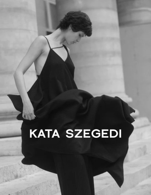 A woman with short dark hair wearing a black dress stands on stone steps in a black and white outdoor fashion shot.