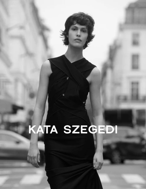 Fashion editorial shot of a woman in a black dress standing on a crosswalk in a city street with buildings and cars in the background.
