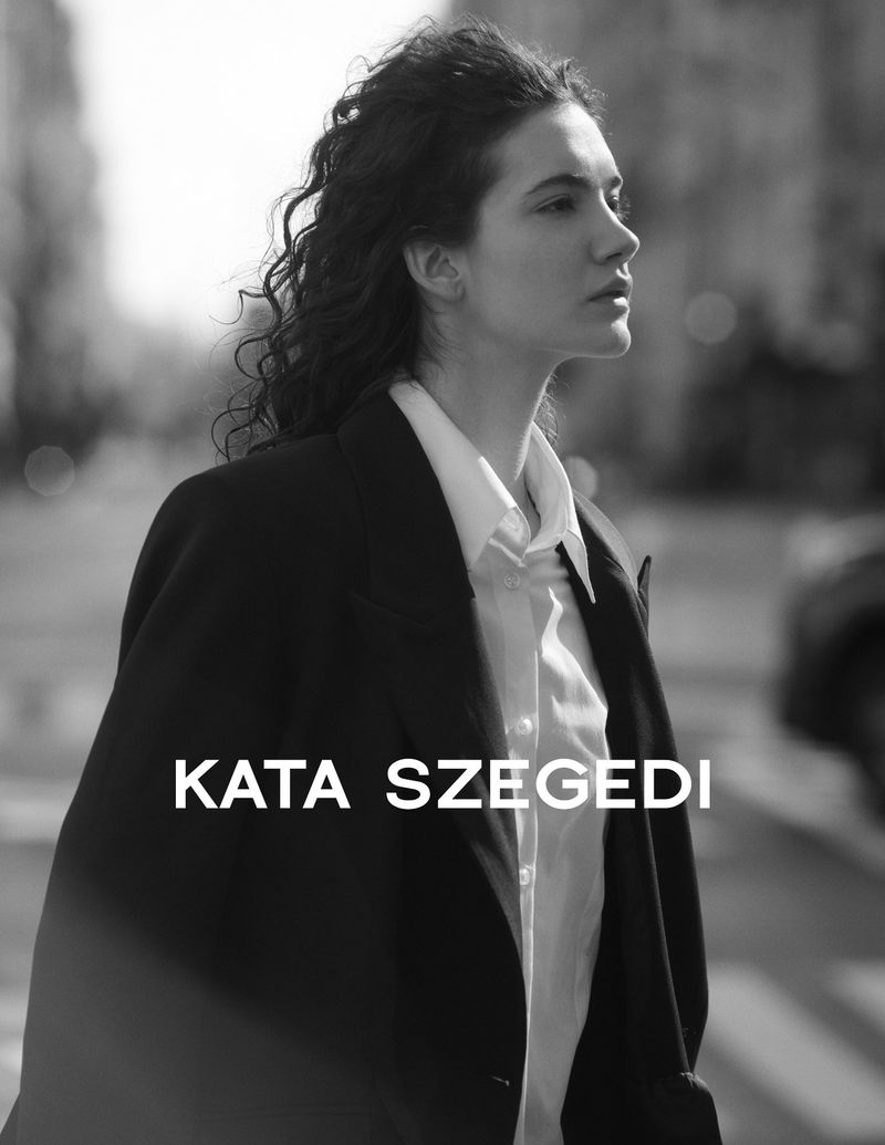 Portrait of a brunette woman with curly hair in a black suit and white shirt, posing outdoors in an urban environment in black and white.