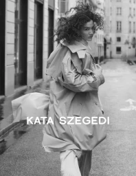 Model with curly hair wearing a belted trench coat walking down an urban street in a black and white fashion editorial image.