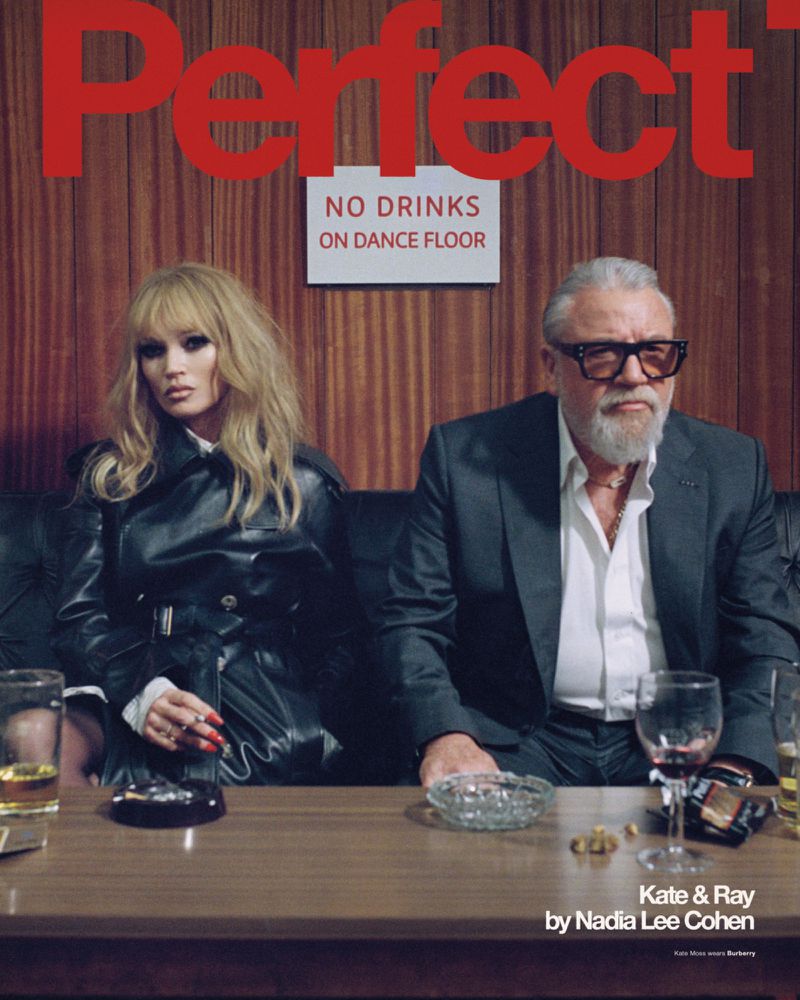 Editorial shot of a blonde woman in a leather jacket and an older man in a suit sitting at a table with drinks in an indoor environment.