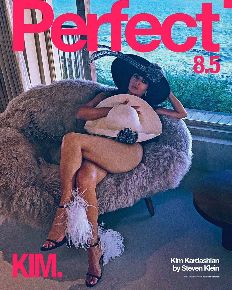 Magazine cover featuring Kim Kardashian in a wide-brimmed hat and feathered heels, seated in a furry chair with an ocean backdrop.