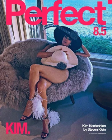 Kim Kardashian in a large feathered hat lounging on a textured grey armchair by a coastal window for The Perfect Magazine.