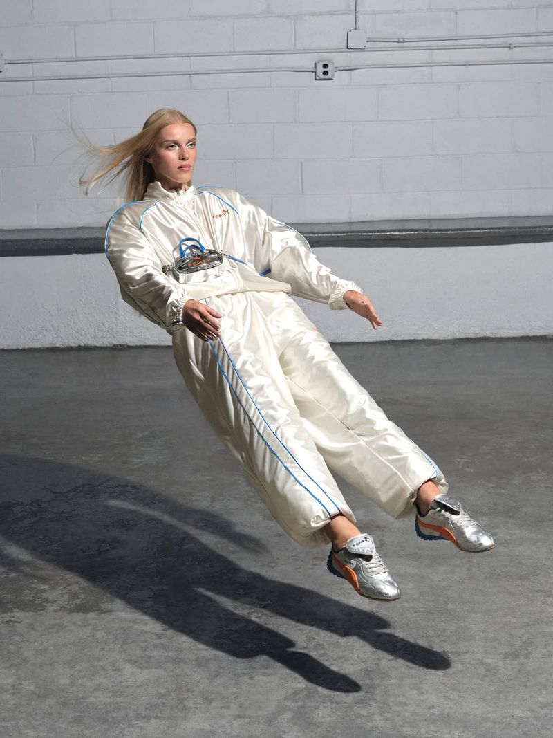 A woman in a white tracksuit and metallic silver sneakers appears to float in a minimalist industrial photo studio against a white brick wall.