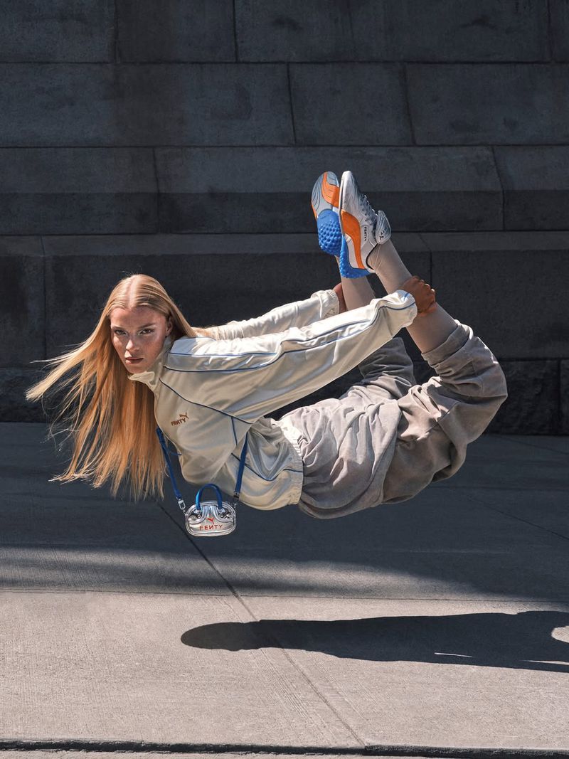Athletic woman wearing a tracksuit and sneakers suspended mid-air in an outdoor urban setting with a grey stone wall backdrop.