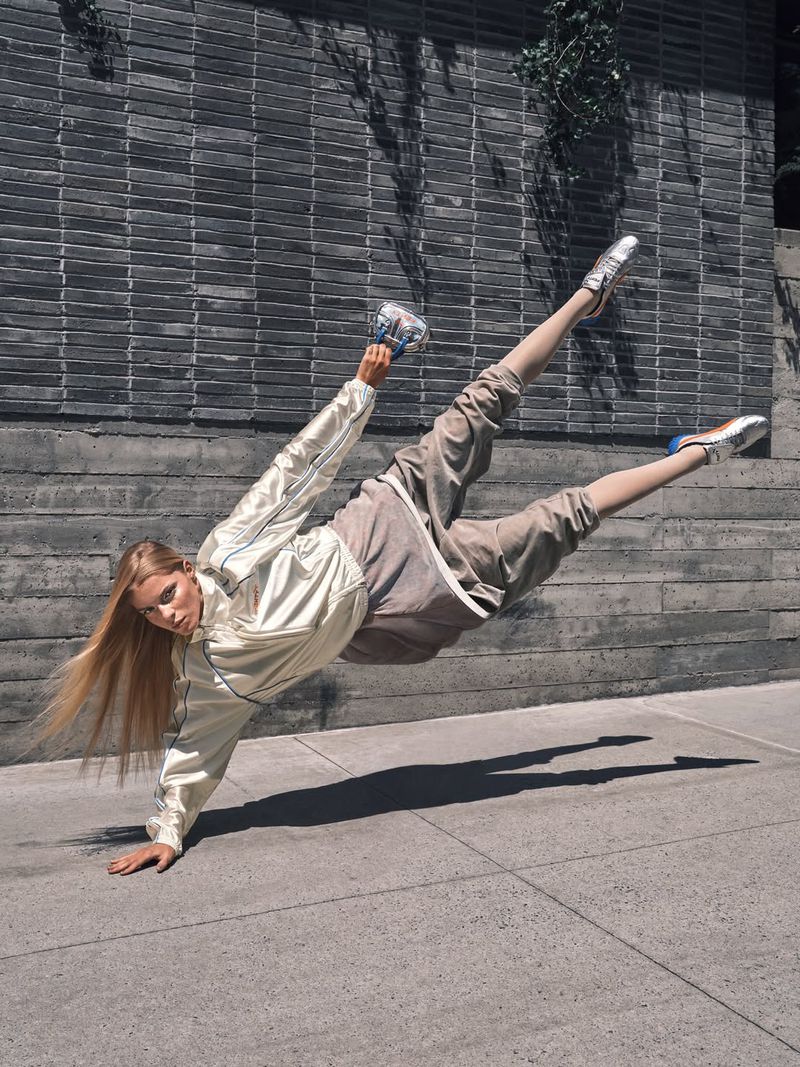 A person in a light-colored jacket and pants performs a dynamic handstand in front of a concrete wall with green foliage.