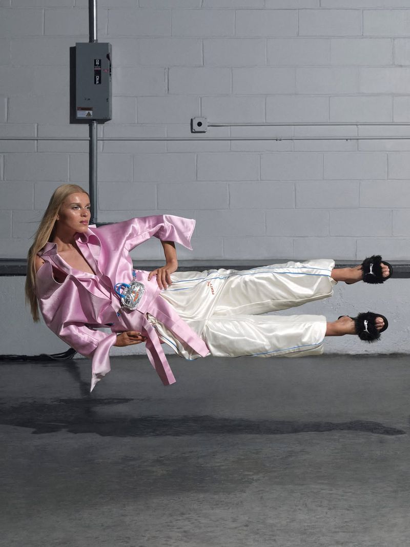 Fashion shot of a woman floating in the air wearing a pink satin robe and white pants, indoors against a grey brick wall backdrop.
