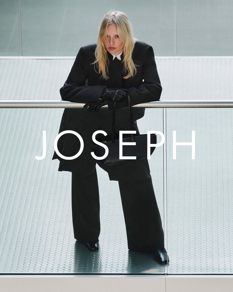 A slender adult woman is wearing a black suit with white shirt and posing with a black purse inside a modern building with glass walls.
