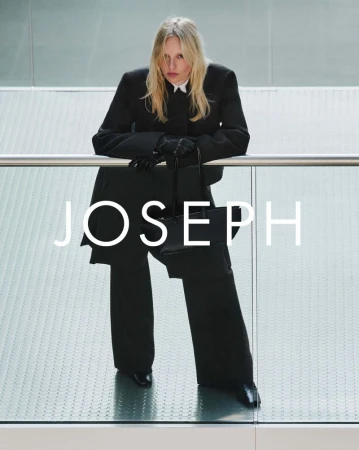 A slender adult woman is wearing a black suit with white shirt and posing with a black purse inside a modern building with glass walls.