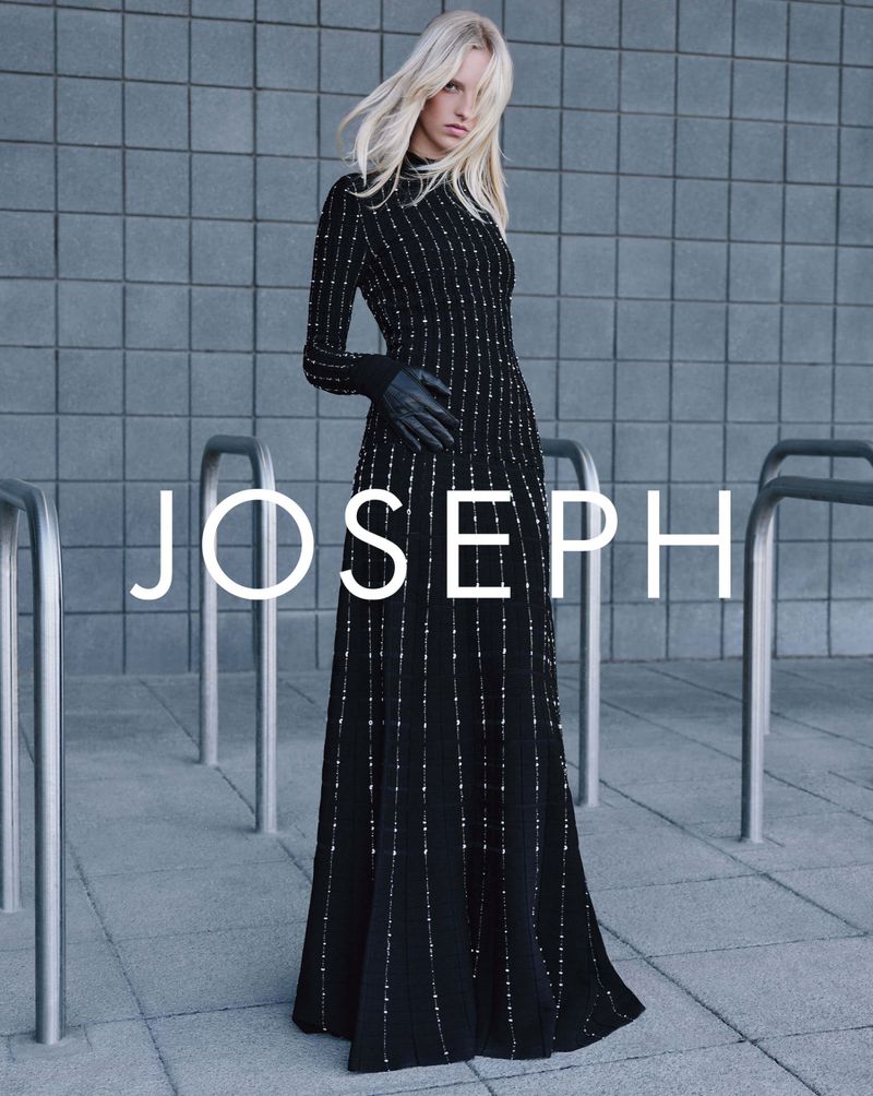 A blonde-haired woman in a black, beaded gown poses outdoors in front of a grey tiled wall and a silver metal structure in a fashion shot.
