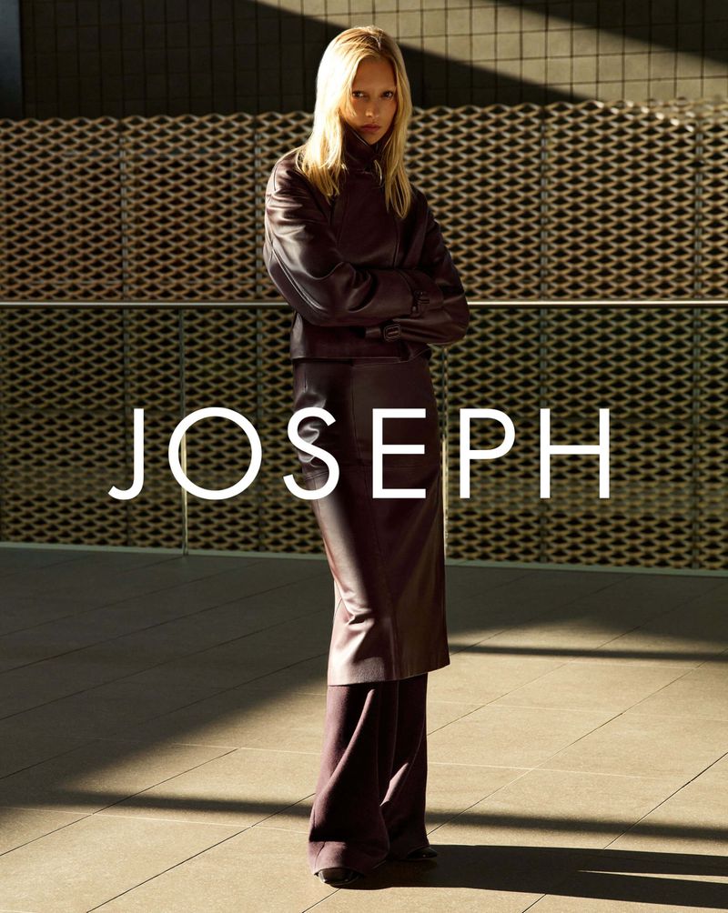 Woman in plum jacket and skirt poses against geometric backdrop; light illuminates Joseph logo; fashion photography, indoor setting.