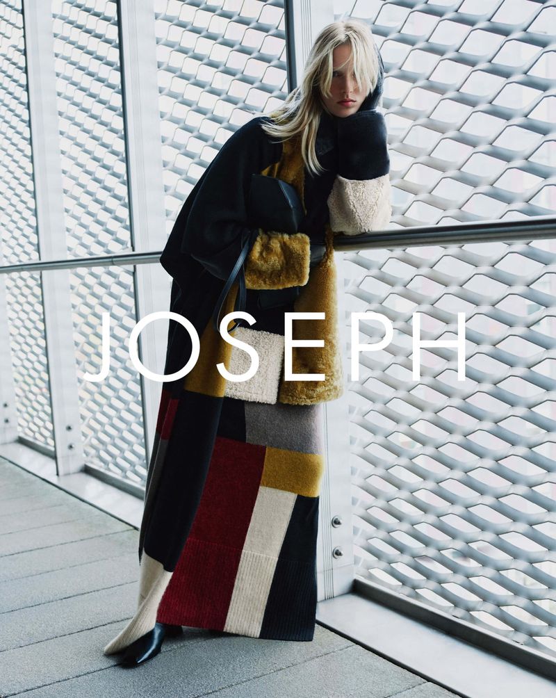 Fashion shot: Blonde woman in black coat, colorful maxi skirt poses against modern geometric background, promoting the brand JOSEPH.