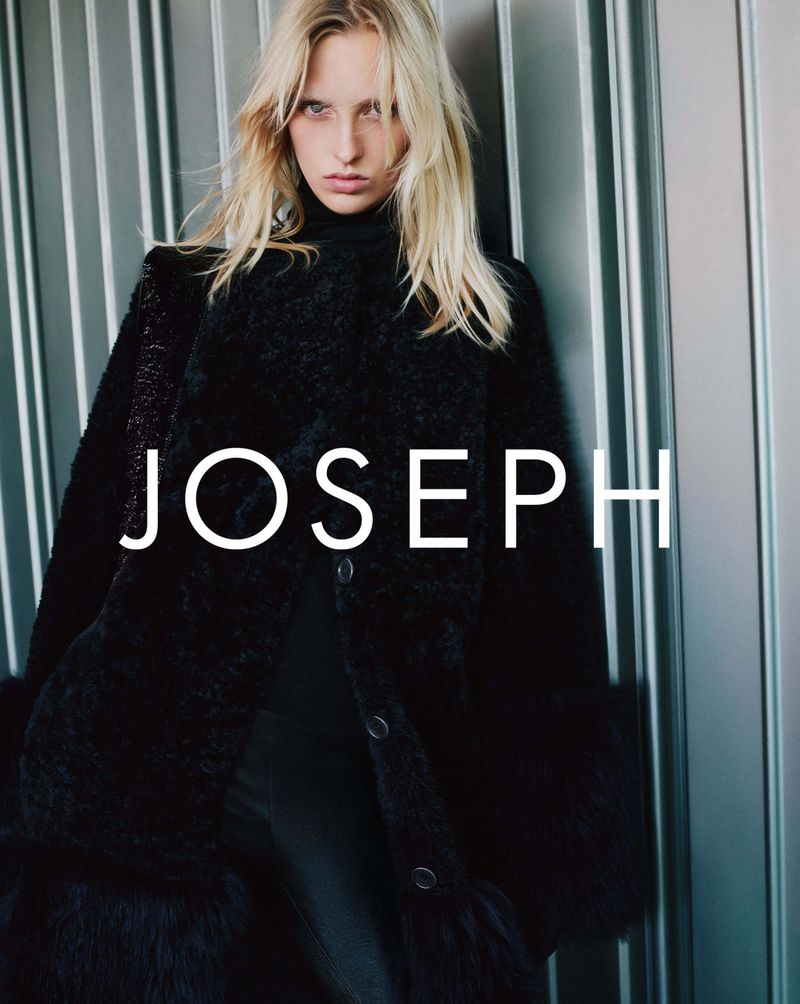 Fashion shot of a blonde adult woman wearing a black coat. The background consists of silver metal panels. The brand name JOSEPH is visible.
