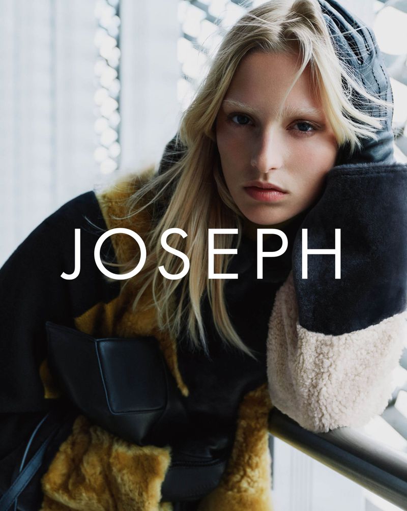 Fashion shot: A fair-skinned fashion model poses in a black coat with fur trim and hood against a blurred, indoor background.