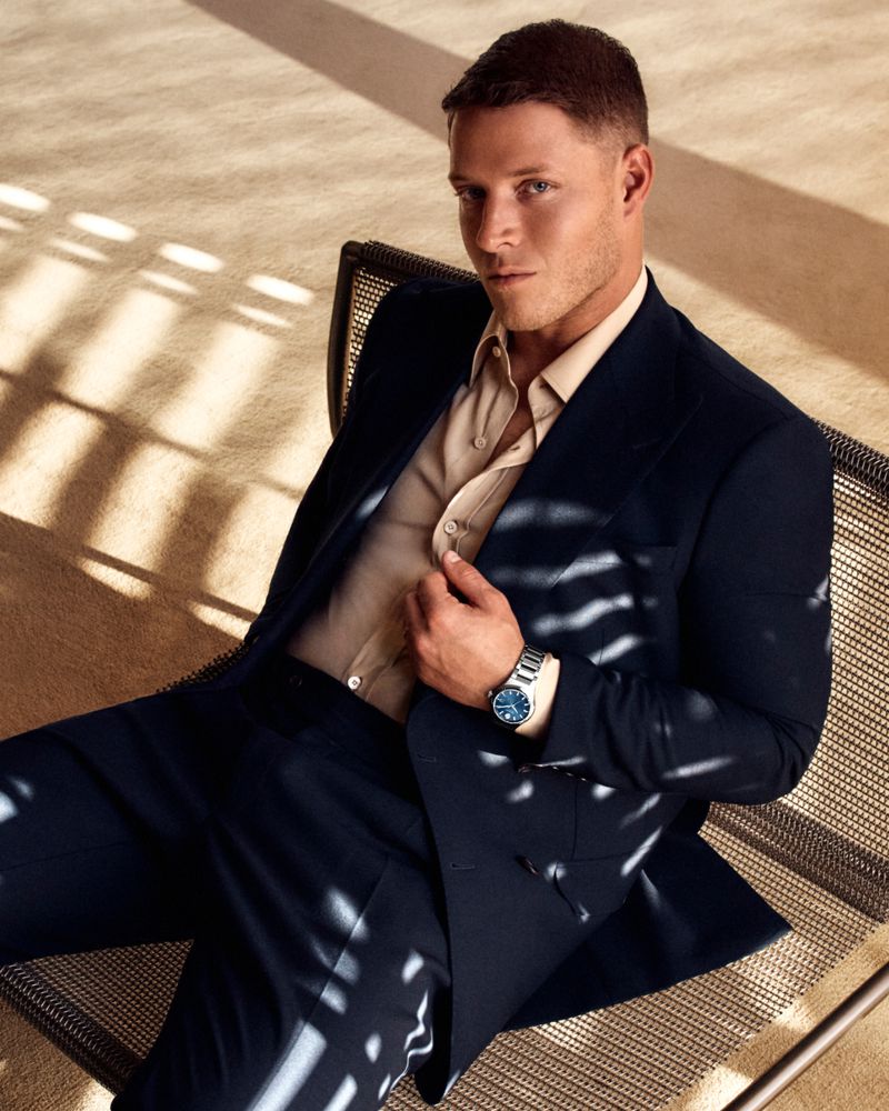 Portrait of a Caucasian adult male in a navy suit and light button-down shirt, seated in a chair with shadow patterns on the floor.