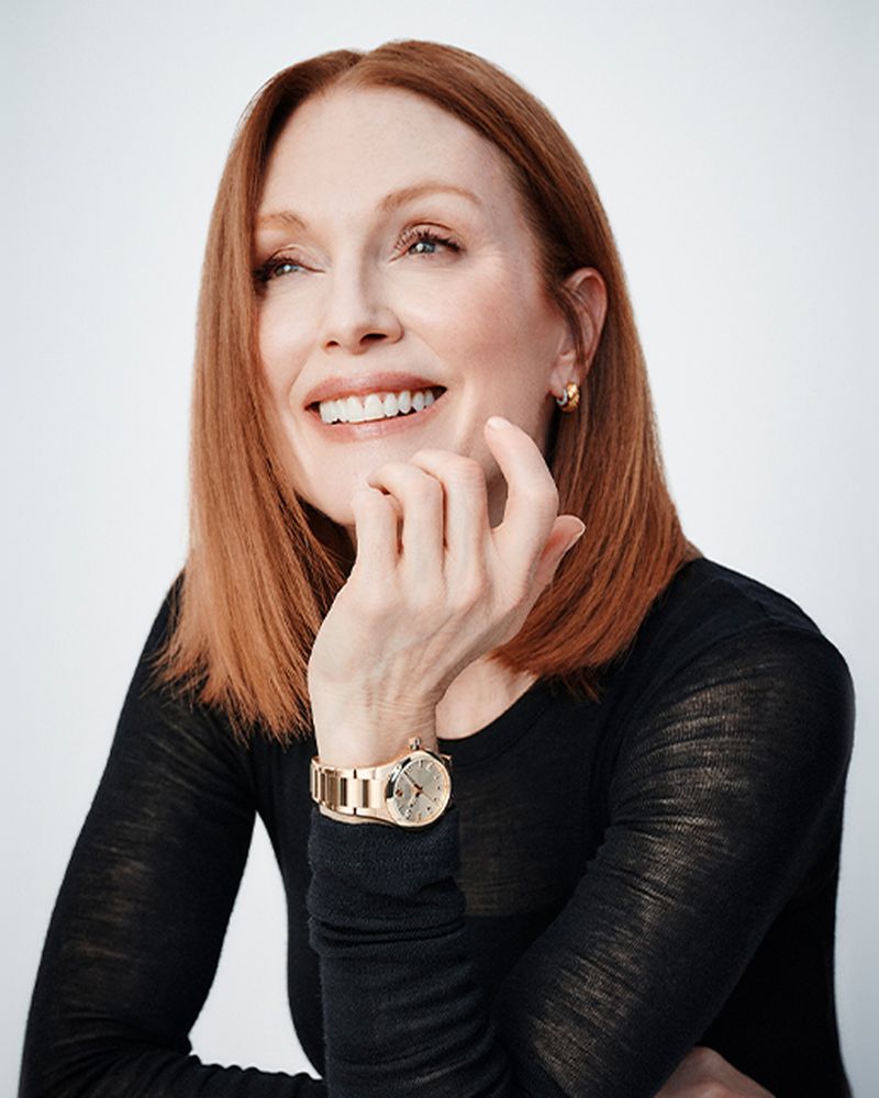 Close-up portrait of smiling adult woman with red hair, wearing a black top and gold watch, against a bright neutral background.