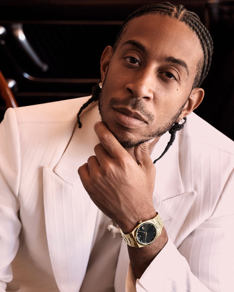 Close-up portrait of an adult male with cornrow braids, wearing a white suit and gold wristwatch, resting his chin on his hand.