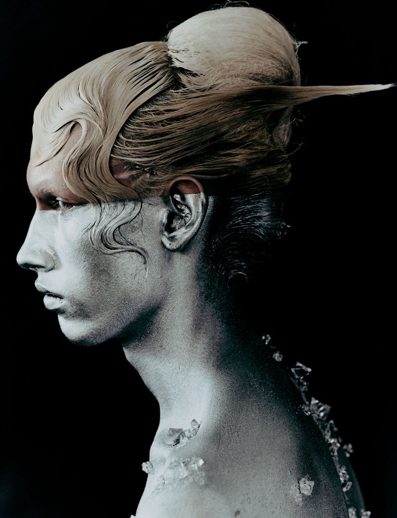 Profile portrait of a person with silver body paint, blonde hair styled up, and crystal shards on their shoulder against a black backdrop.