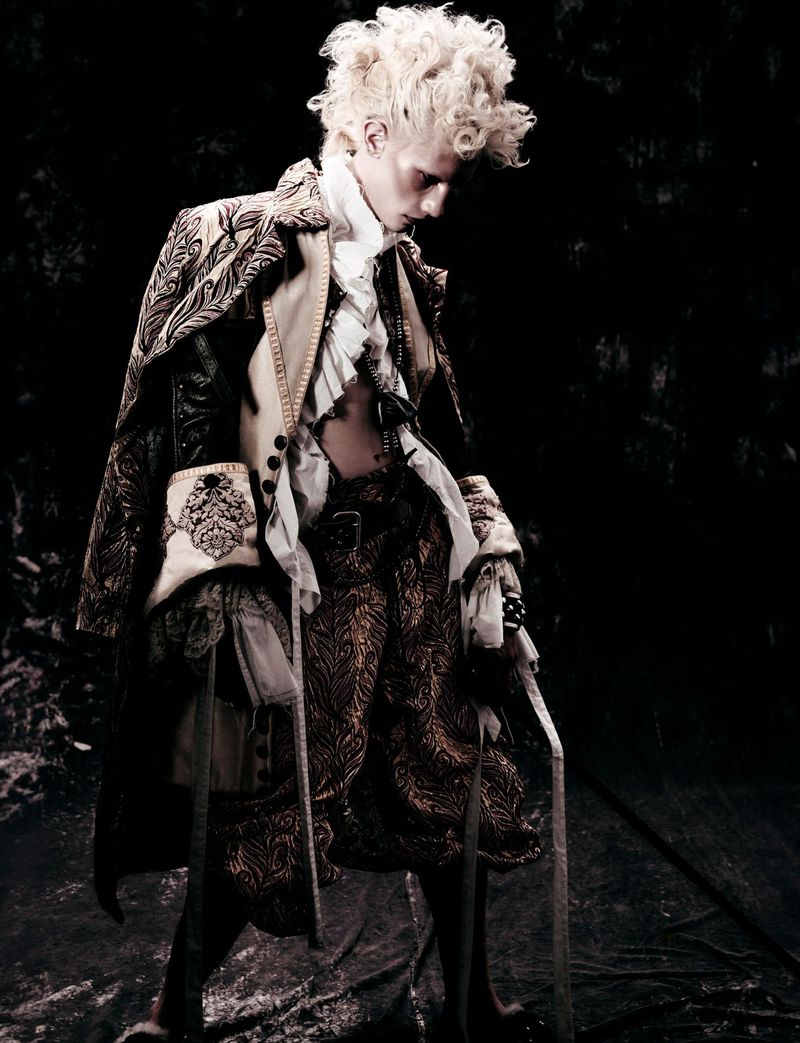 A person in an ornate, vintage-style costume stands against a dark background, exhibiting intricate fabric details and period fashion.