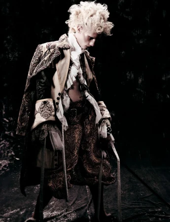 A person in an ornate, vintage-style costume stands against a dark background, exhibiting intricate fabric details and period fashion.