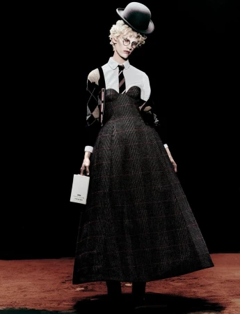 Fashion model in bowler hat and gray plaid dress, posing in a studio setting against a dark background, holding a white box.