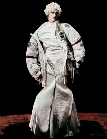 Fashion model with curly blonde hair wears white space suit dress with NASA logo and American flag against black and brown backdrop.