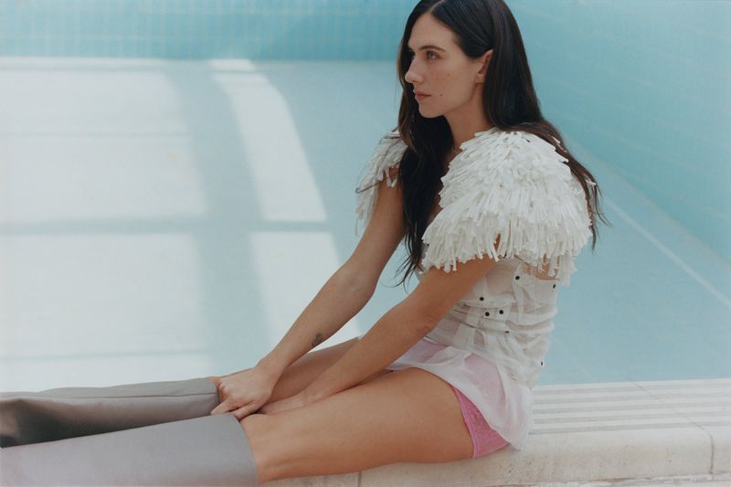 A woman with long dark hair poses seated on a ledge wearing a white fringed top and pink bottoms in front of a light blue tile wall.