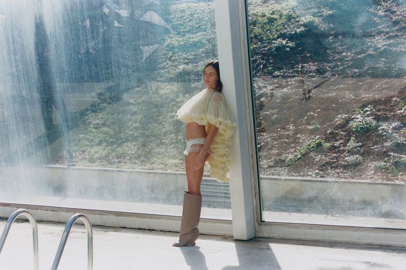 Woman in voluminous yellow tulle top and thigh-high boots poses against a glass wall near a pool in natural light.