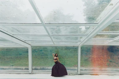 Devon Lee Carlson in a dark two-piece outfit standing inside a glass-roofed pavilion for a Russh editorial by Tatiana and Karol