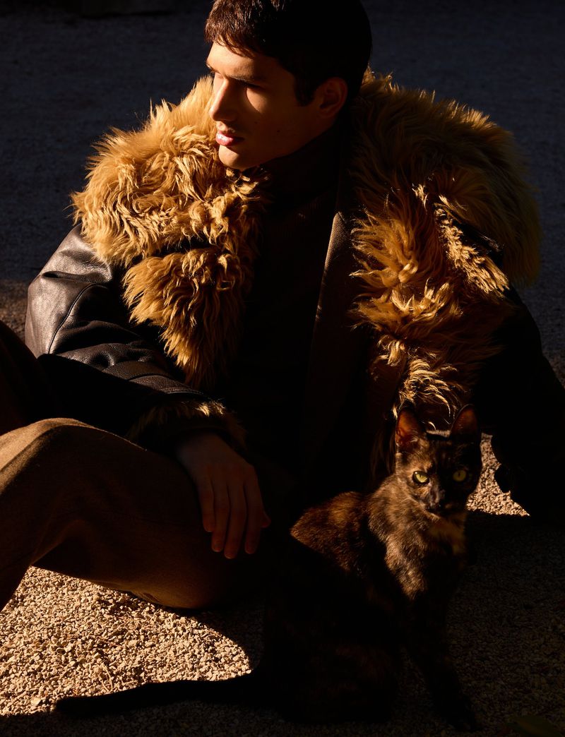 Model Luka Tesic wearing a dark leather jacket with a voluminous shearling collar, seated outdoors next to a cat in bright sunlight.