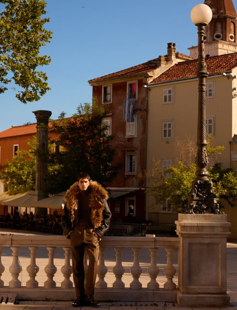 Adult male in brown fur collar coat poses on marble railing with old buildings in background, lit by warm sunlight in an outdoor urban setting.