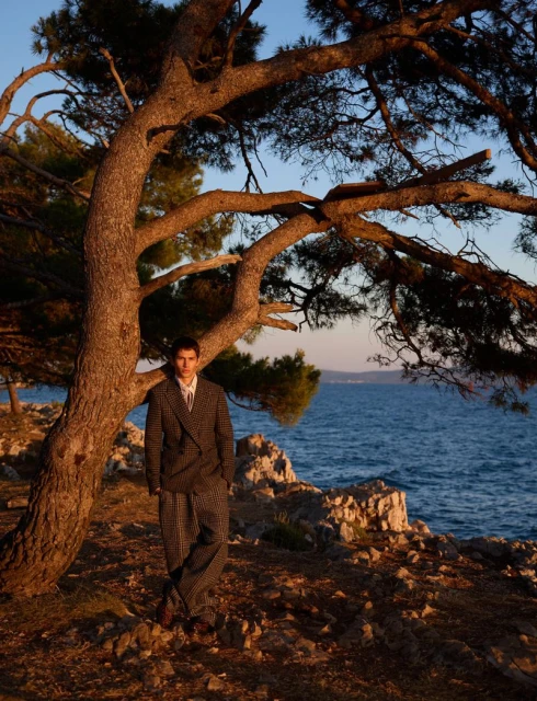 A male model poses in a brown checkered suit under a large pine tree overlooking the ocean at sunset; rocks, blue water and clear sky.