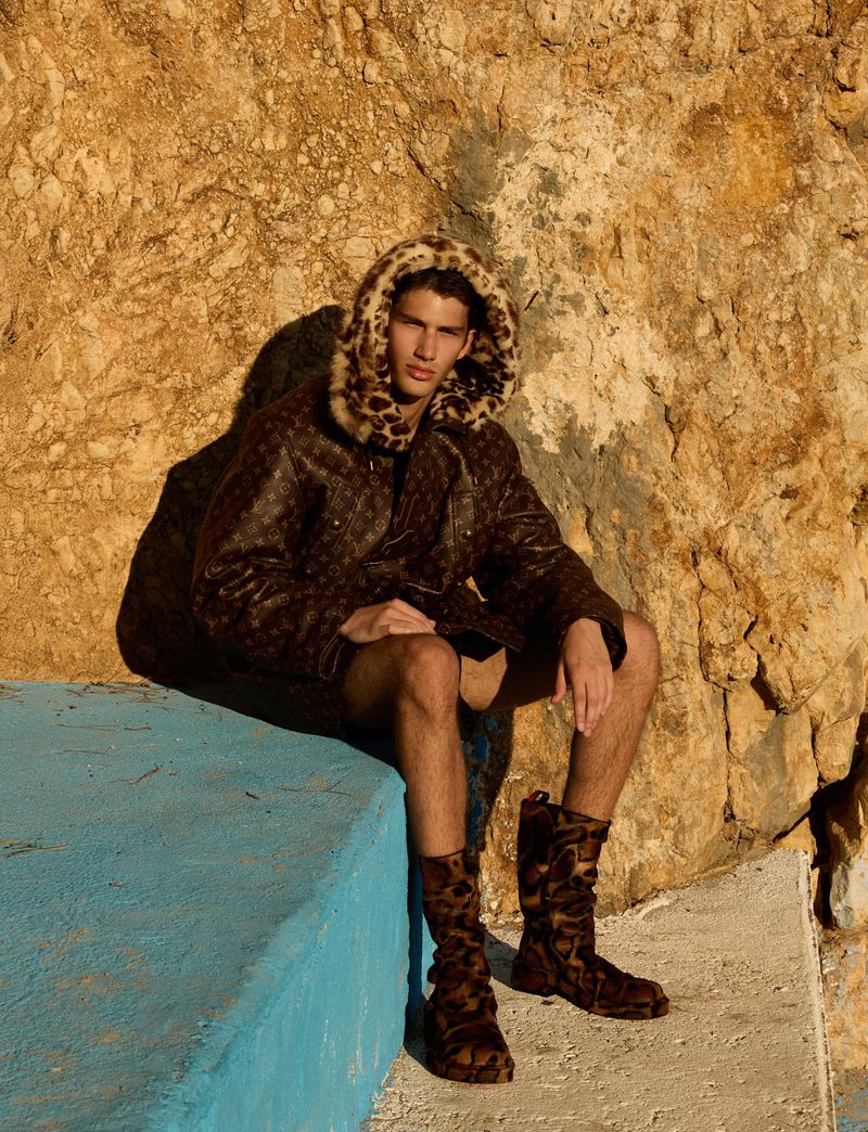 Male model in patterned jacket and boots poses outdoors against a rocky wall and teal ledge, casting a shadow in warm light.