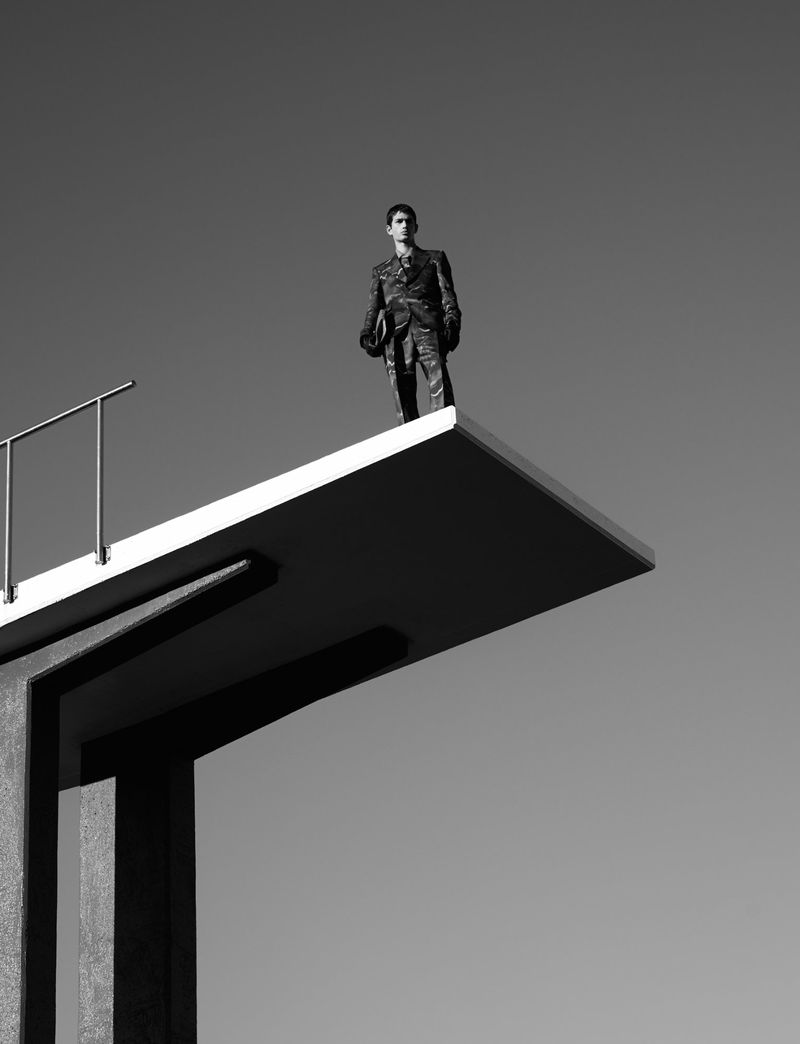 Man in a suit stands on a diving platform against a clear sky in a black and white photograph; the platform juts out over the frame.
