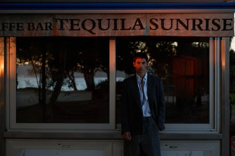 Young man in a suit poses in front of a vintage cafe bar with ocean views in the background at golden hour, fashion editorial photography.