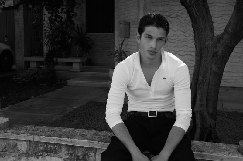Black and white portrait of model Luka Tesic wearing a white long-sleeve polo shirt and dark trousers outdoors on a stone wall.