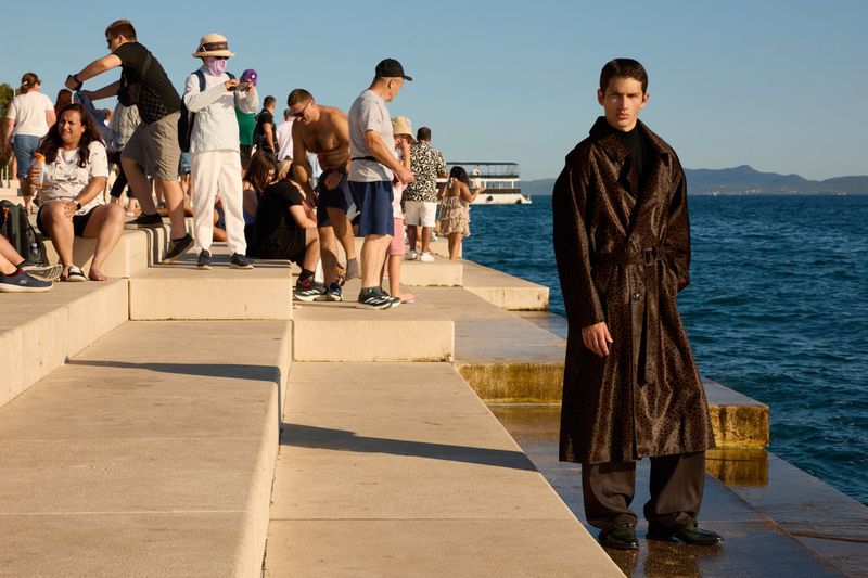 Man in patterned brown coat stands on a concrete pier with a group of people and the sea in the background on a sunny day.