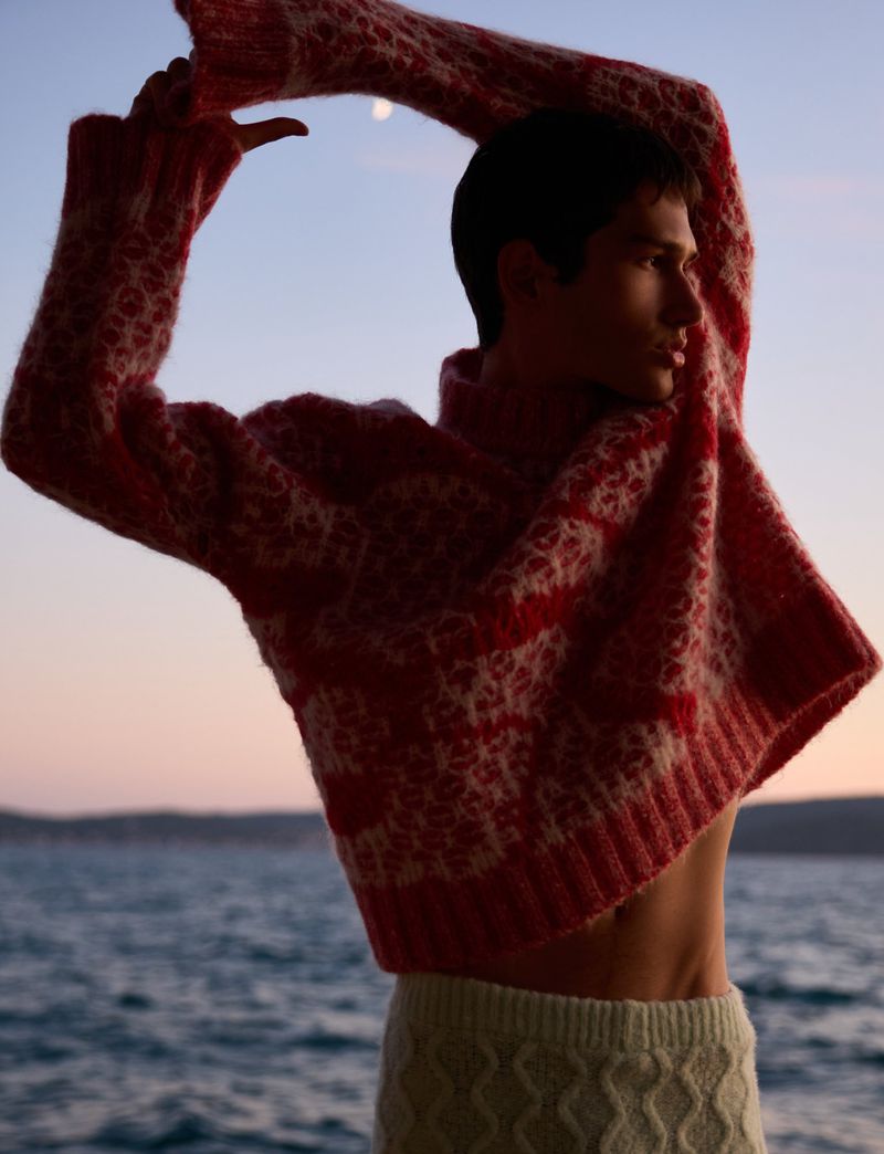 A slender man in a red knit sweater and white knit shorts poses outdoors near water at dusk, showcasing fashion and knitwear.