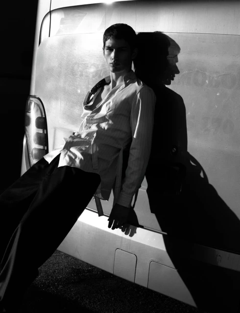 A young adult man in an unbuttoned shirt and dark pants leans against a metallic van with a dark shadow in a monochrome shot.