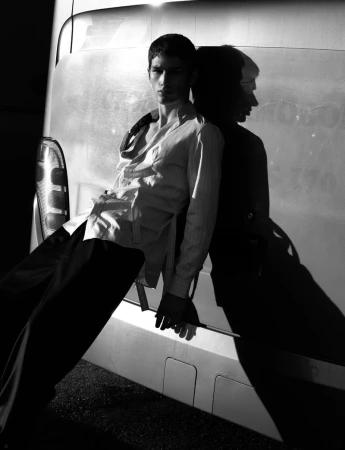 A young adult man in an unbuttoned shirt and dark pants leans against a metallic van with a dark shadow in a monochrome shot.